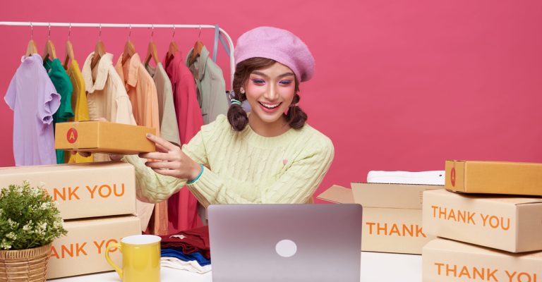 Young asian woman startup small business freelance sale fashion clothing with parcel box and computer laptop on table and sitting isolated on pink background. Online marketing and delivery concept.